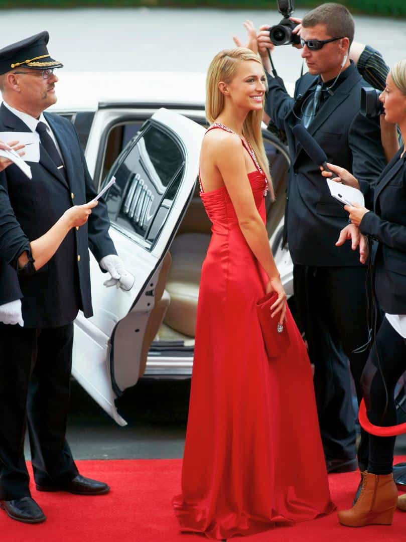 Celebrity walking red carpet in red dress while paparazzi and reporters surround h